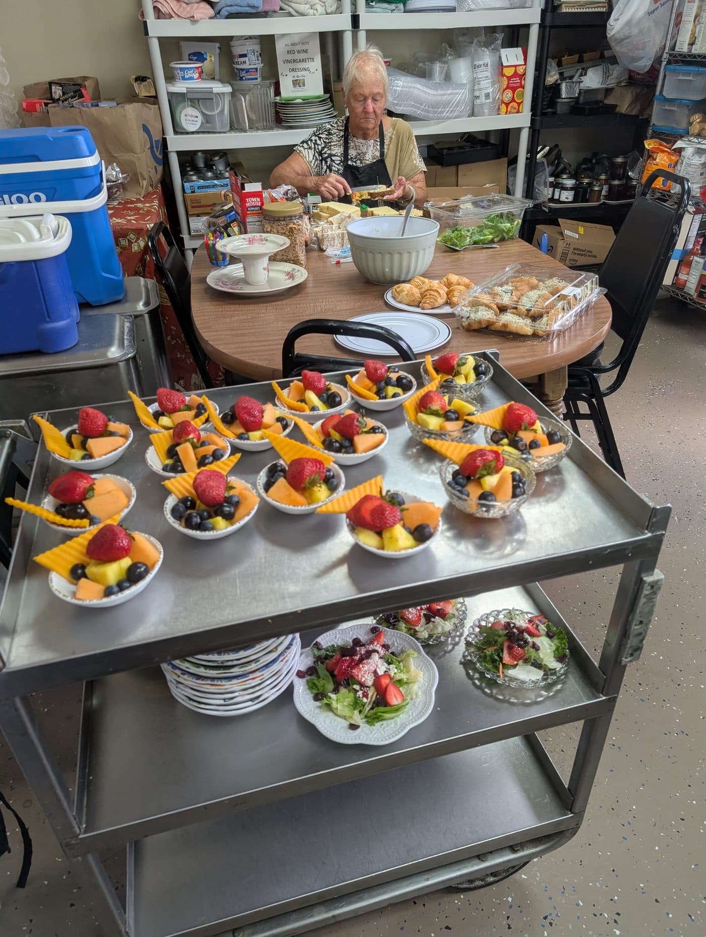 Ginny in her apron making sandwiches, trays of fruit cups ready on the cart